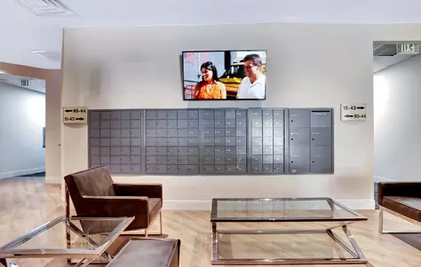 A wall of mailboxes with a flat-screen TV mounted above them, with cushioned chairs and glass coffee tables occupying the open space
